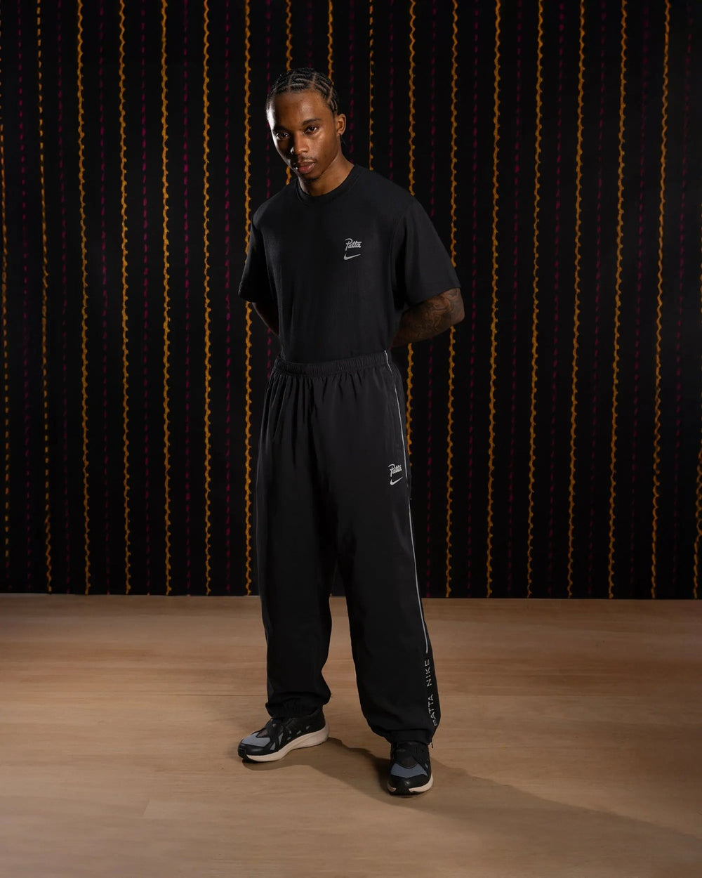 Model wearing black Nike x Patta Running Team track pants with reflective piping and logo details.
